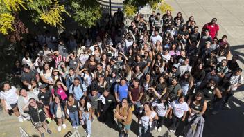A large group of students gathers outdoors in a shaded courtyard, looking up at the camera. Some students wear 'College Corps' shirts, indicating their affiliation with the program. The crowd is smiling, with name tags visible on some attendees. Trees with green and yellow leaves partially frame the scene.