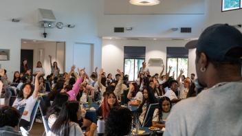 A large group of students is gathered in a bright event space, many raising their hands in response to a speaker at the front. The speaker, wearing a cap, addresses the engaged audience. Some attendees wear shirts with the 'College Corps' logo, indicating a College Corps-related event.