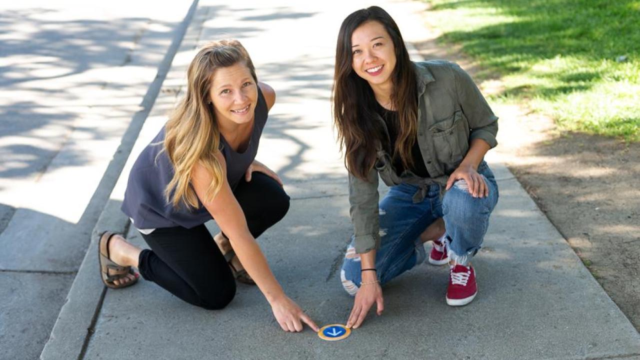 Two women point to walking loop sign on sidewalk.