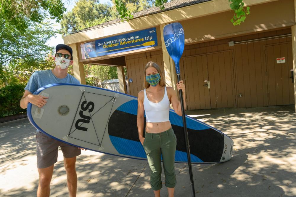 Two students stand outside Outdoor Adventures, holding a paddleboard and paddle.